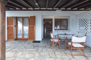 Une terrasse dallée de pierres, avec une table ronde et des chaises blanches, des volets en bois et un treillage.