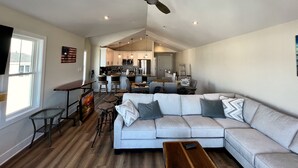A spacious living room with a large sofa, a coffee table, and a dining area with a kitchen in the background. - Stunning  Townhome, Private Elevator, Expansive Bay Views! (Sea Isle City)