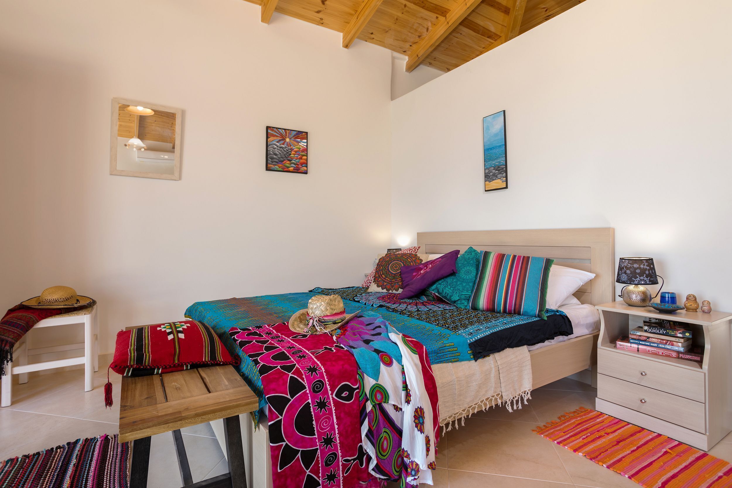 Een slaapkamer met een houten hoofdbord, een nachtkastje met een lamp, een klein plankje met boeken en een kleurrijk vloerkleed.