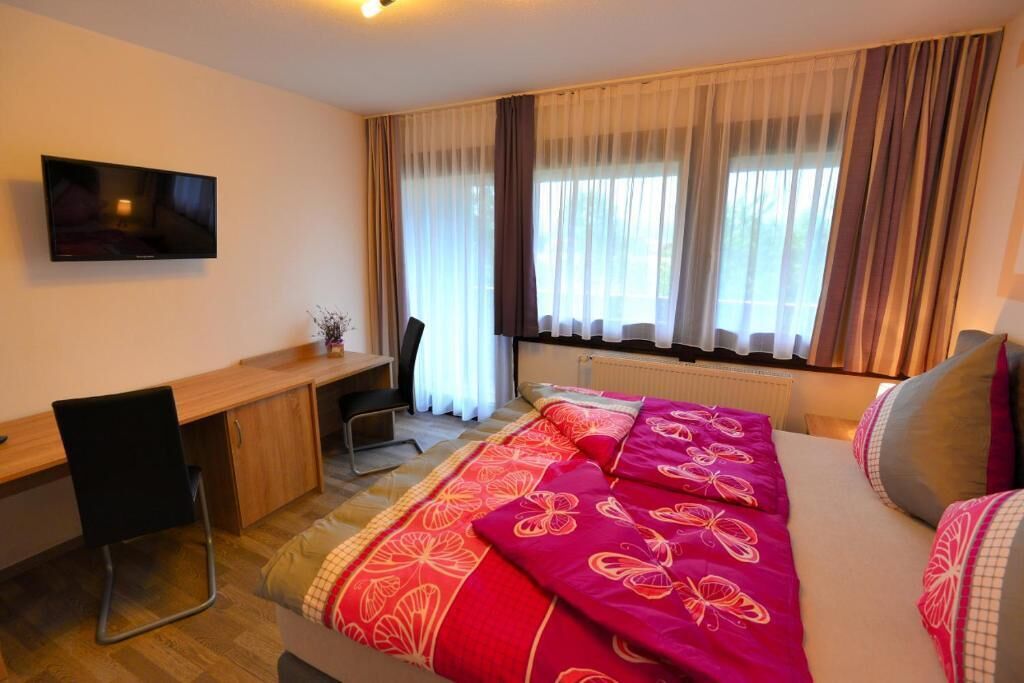 Una habitación de hotel con cama, escritorio, silla, televisión y ventana con cortinas.