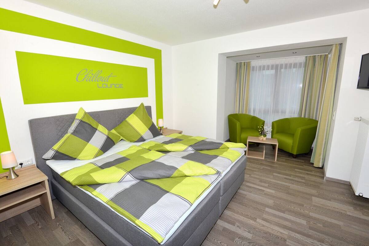A modern hotel room with a large bed, a bedside table, a green armchair, and a view of the outdoors through a sliding door.