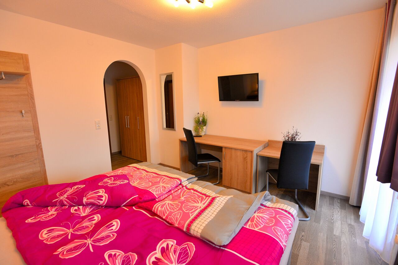 A bedroom with a bed, a desk, a chair, and a TV.