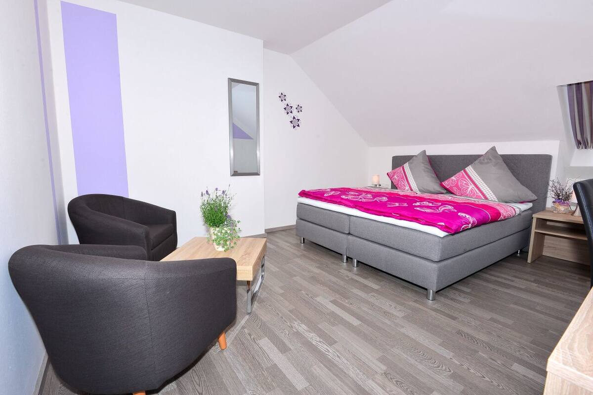 A modern bedroom with a grey sofa, a wooden side table, a bed with a pink and grey patterned bedspread, and a small potted plant.