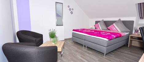 Un dormitorio moderno con un sofá gris, una mesita de madera, una cama con colcha estampada en rosa y gris, y una pequeña planta en maceta.