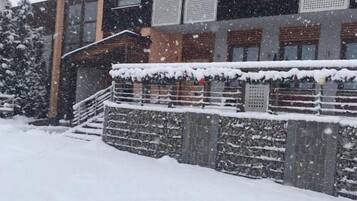 A multi-story building with a stone facade and multiple windows. Snow covers the ground and the roof.