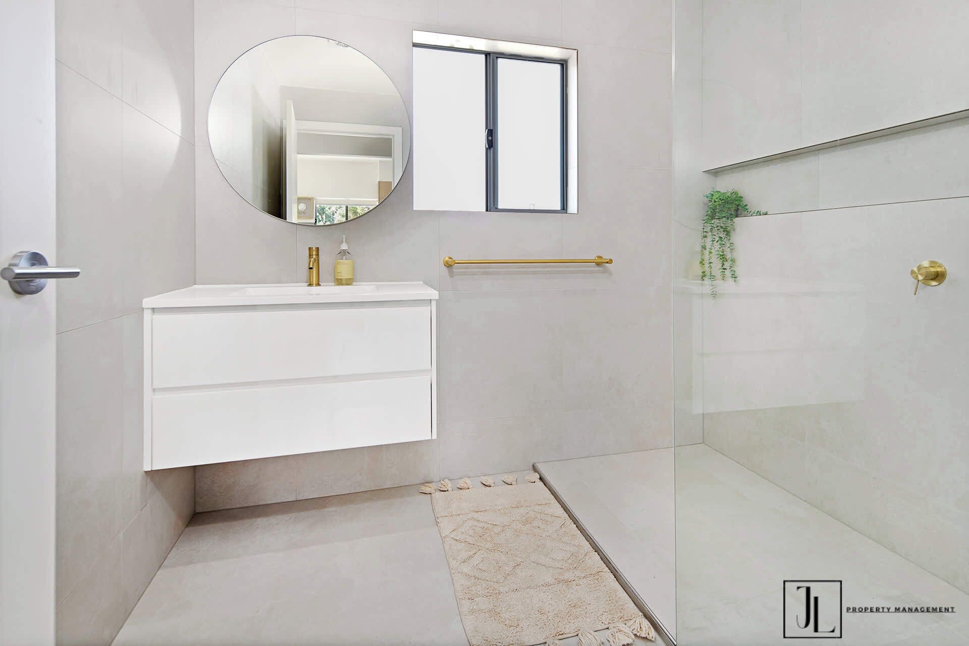 Un baño moderno con un tocador blanco, un espejo redondo, una cabina de ducha de vidrio y una planta pequeña en la pared.