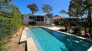 A modern house with a swimming pool, surrounded by tropical plants and a clear blue sky.