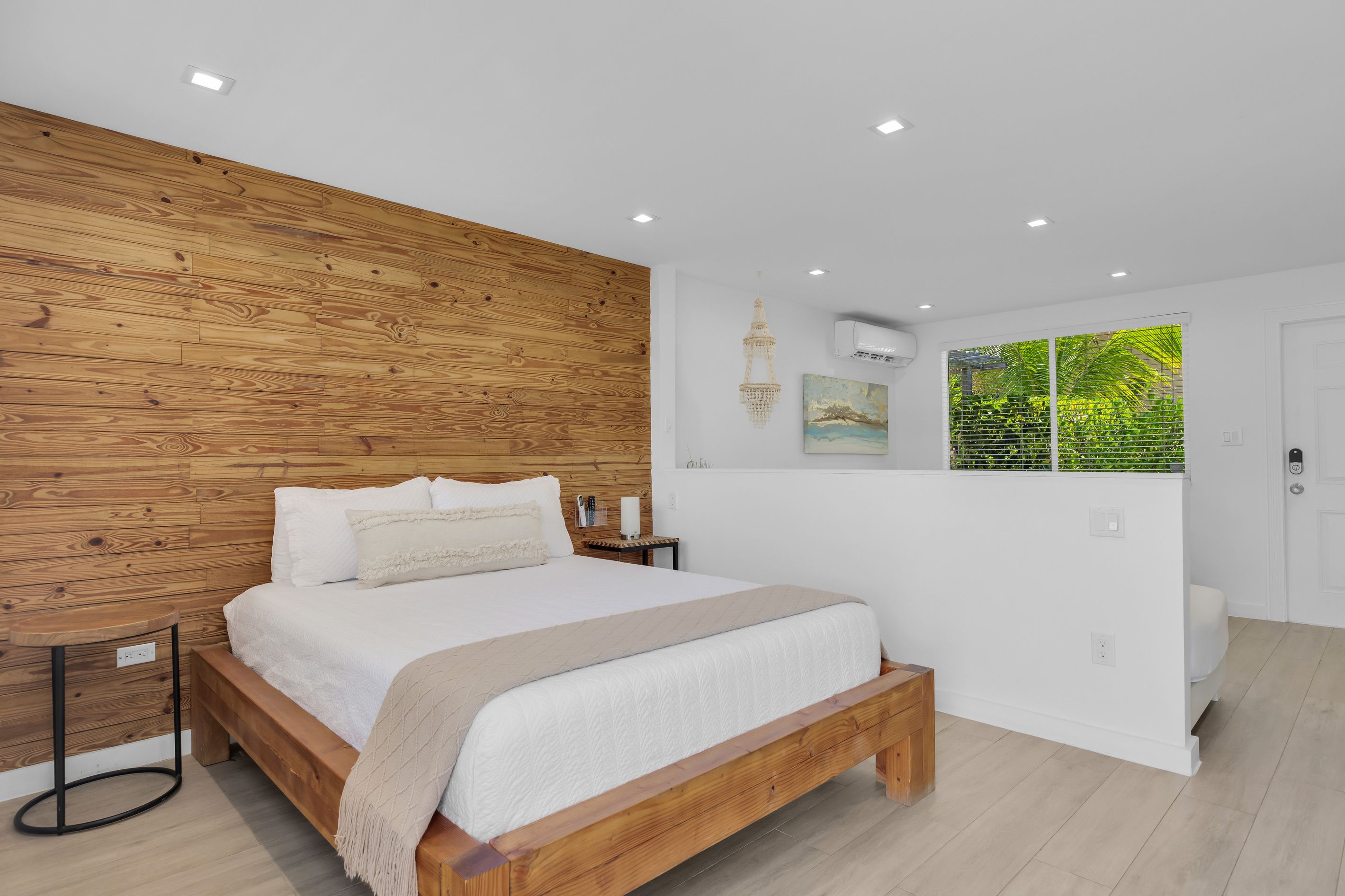 Een slaapkamer met een houten hoofdbord, een bed met wit beddengoed, een nachtkastje, een hanglamp en een raam met uitzicht op groen.