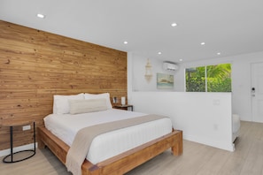 A bedroom with a wooden headboard, a bed with white bedding, a bedside table, a hanging light fixture, and a window with a view of greenery.