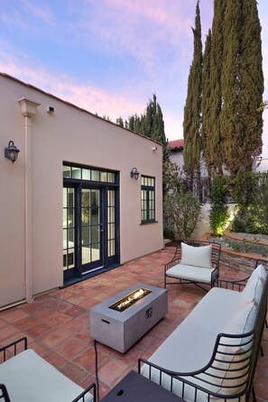 Un patio con mobiliario exterior, una chimenea y un edificio con ventanas y puertas grandes.