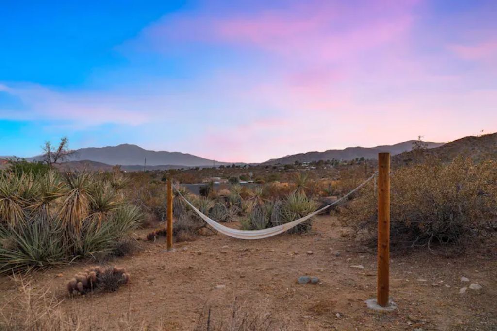 A desert landscape with a hammock tied between wooden posts, surrounded by dry vegetation and mountains in the distance.