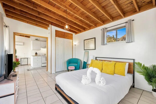 Un dormitorio con techo de madera, una cama con ropa de cama blanca y almohadas amarillas, un sillón turquesa, y vista al exterior a través de una ventana.