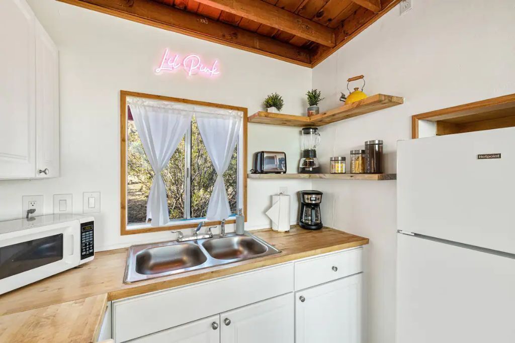 A modern kitchen with a neon sign, a window with curtains, a coffee maker, a toaster, and a refrigerator.