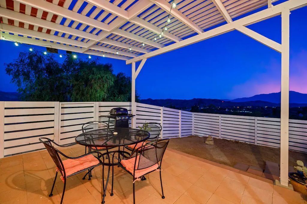 An outdoor patio with a round table and chairs, a grill, and a view of the mountains at dusk.