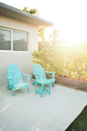 Twee lichtblauwe Adirondack-stoelen op een betonnen terras met uitzicht op een huis en tropische begroeiing.
