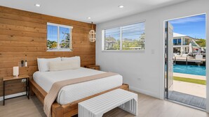 A bedroom with a wooden headboard, a bed with white bedding, a bedside table, and a bench. There are two windows with blinds, a ceiling light, and a view of a pool outside. - 6. Key Largo Waterfront Studio | Dock, Snorkel (Key Largo)