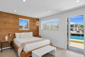 A bedroom with a wooden headboard, a bed with white bedding, a bedside table, and a bench. There are two windows with blinds, a ceiling light, and a view of a pool outside. - 6. Key Largo Waterfront Studio | Dock, Snorkel (Key Largo)