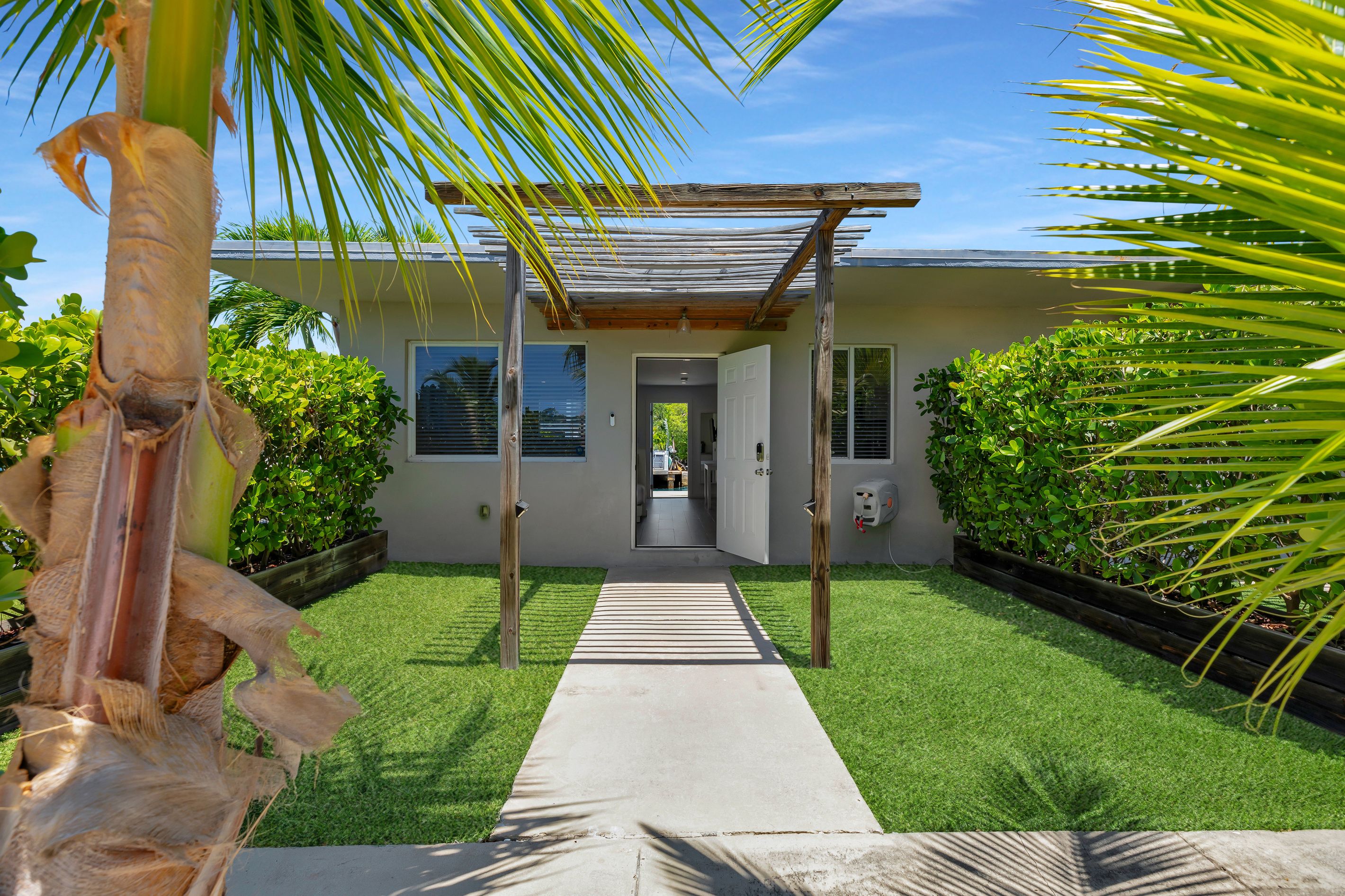 A modern house with a concrete pathway leading to the entrance, surrounded by lush greenery and a wooden pergola.