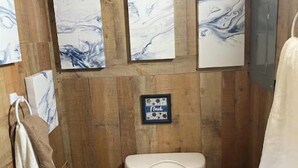 A wooden-paneled bathroom with a toilet, a towel rack, and three marble-patterned wall art pieces. - Delightful kitchenette style tiny home with loft. (Overton)