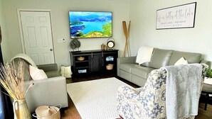 A living room with a grey sofa, a patterned armchair, a coffee table, and a television on a stand. There is a framed picture on the wall and a door in the background. - Quaint 2 bedroom cottage at Bass Lake-paddle boards, kayaks, private ramp, bikes (Knox)