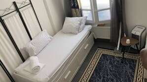 A small, single-bed room with a window, a radiator, a heater, and a small table with a heater.