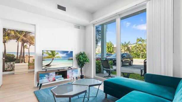 A modern living room with a television, sofa, and a view of a beach and palm trees through large windows. - Stylish & Cozy ~ 15mins to Downtown & Beach ~ Pool (West Palm Beach)