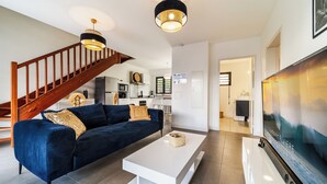 A modern living room with a blue sofa, white coffee table, and a TV. A wooden staircase and kitchen are visible in the background.