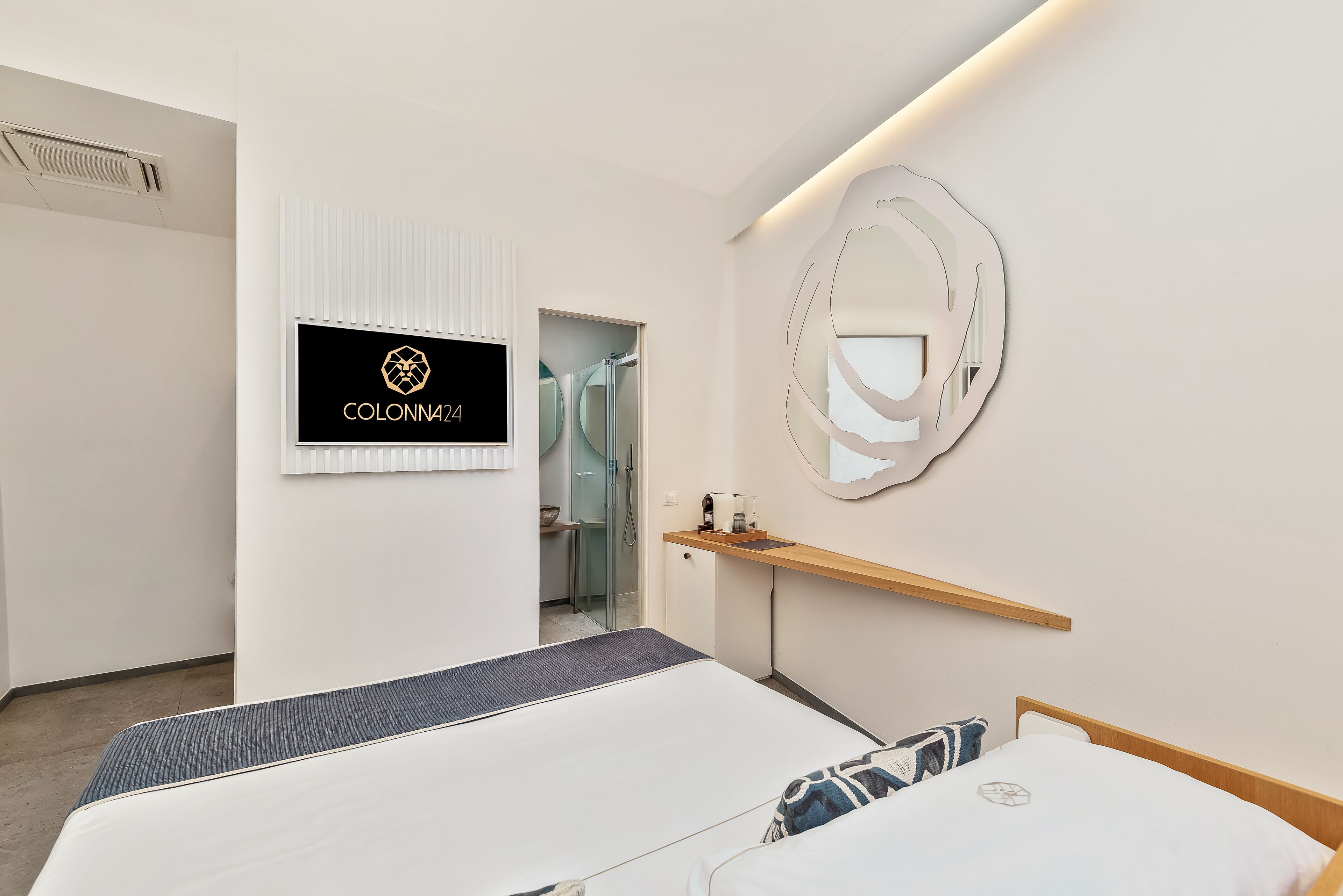 Habitación de hotel moderna con una cama, un tocador con espejo, un estante pequeño y un televisor mostrando 'COLONN424'.