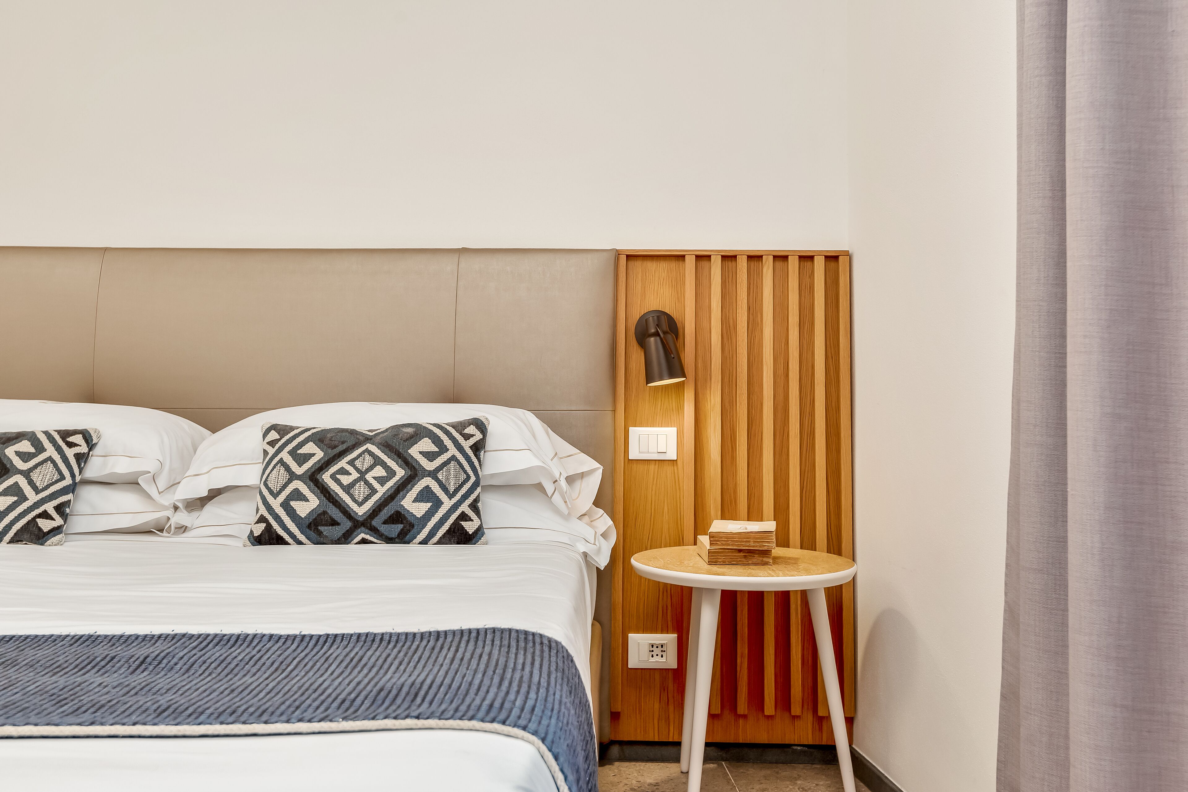 A neatly made bed with patterned pillows, a small round table with a book, and a wall-mounted lamp.