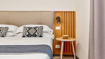 A neatly made bed with patterned pillows, a small round table with a book, and a wall-mounted lamp.