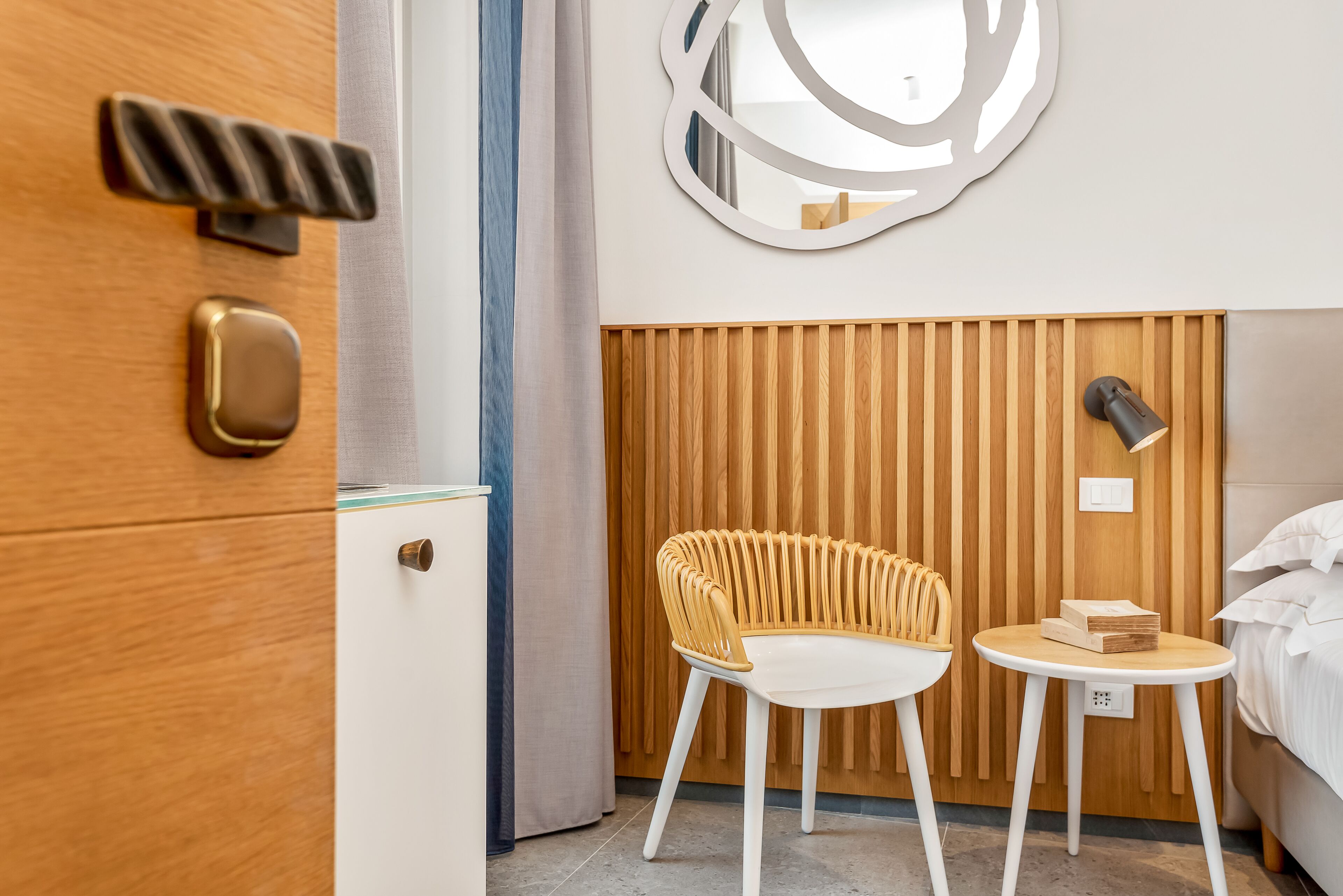 A modern hotel room with a wooden headboard, a round mirror, a wicker chair, a small table with books, and a bed with white linens.
