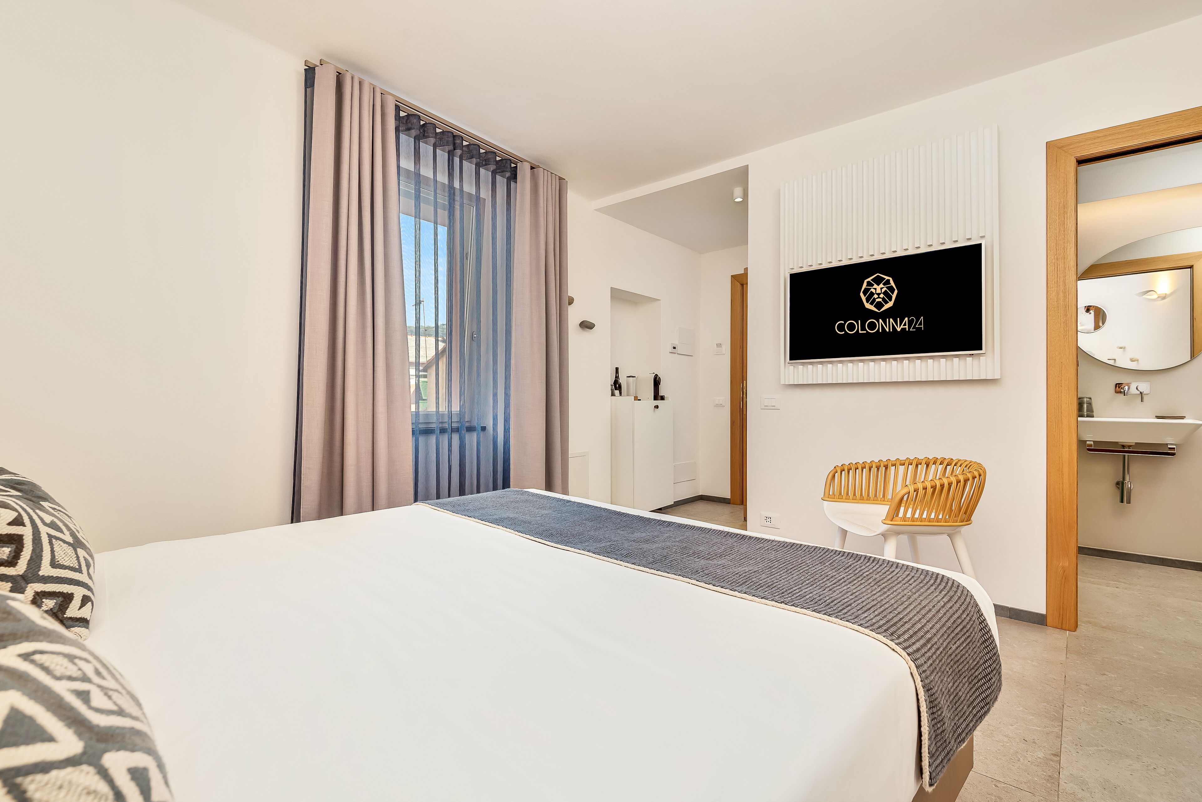 A hotel room with a bed, a TV mounted on the wall, a wooden chair, and a bathroom visible through an open door.