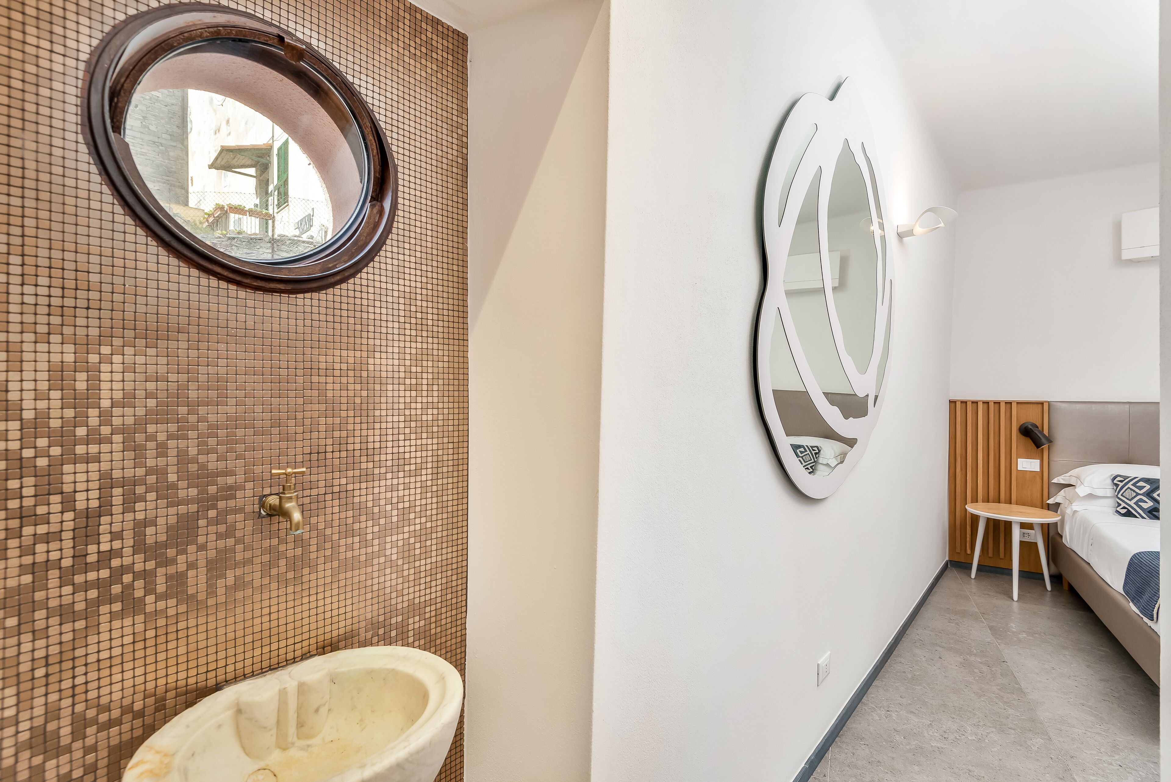 Un bagno con parete rivestita in mosaico, uno specchio rotondo, un lavandino e un letto con testiera in legno.