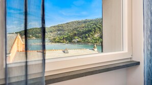 A view from a window overlooking a hillside with a body of water, a building with a sloped roof, and a clear blue sky. - COLONNA24 (Portovenere)