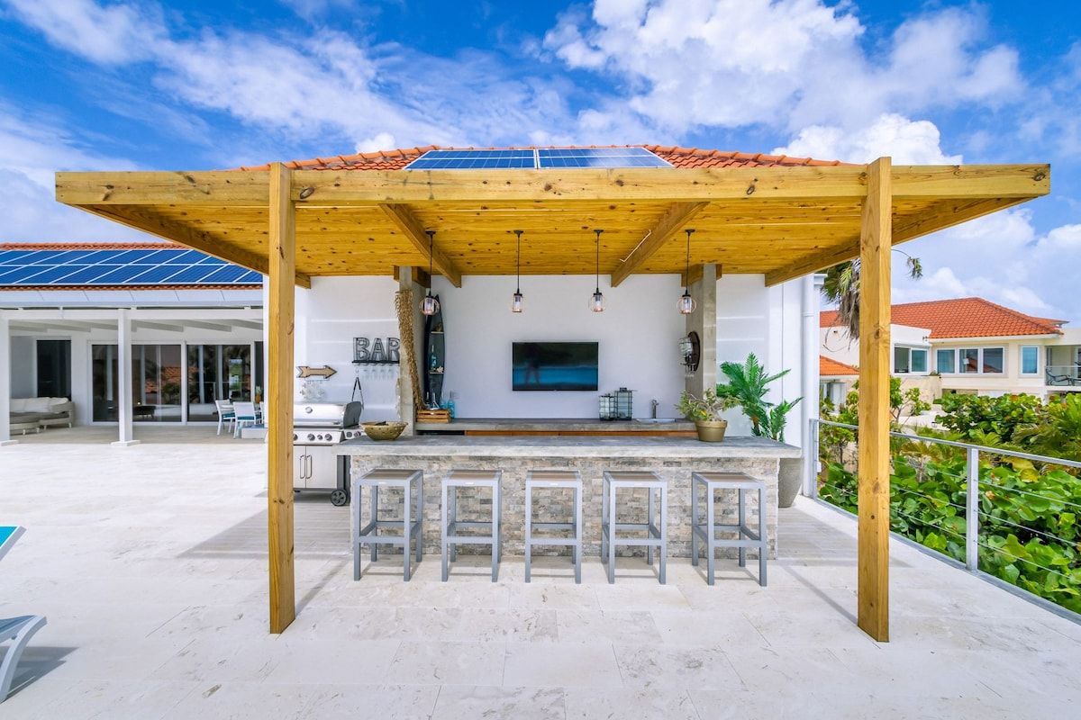 Un espace bar extérieur doté d’une pergola en bois, d’un écran de télévision et de tabourets de bar.