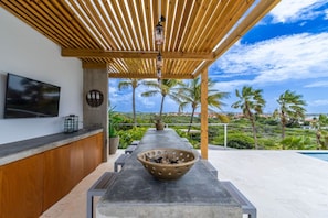 A covered outdoor area with a concrete table, a bowl of fruits, a television, and a view of palm trees and hills.