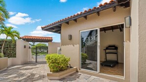 Un gimnasio con puerta de vidrio corredera, una cinta de correr en su interior, y un patio con piso de baldosas y un pequeño arbusto.