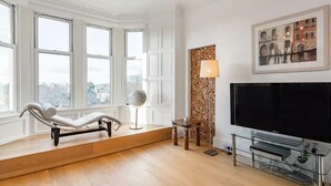 Een moderne woonkamer met een chaise longue, een grote flatscreen-tv en een stadsgezicht door grote ramen.