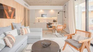A modern living room with a sofa, armchair, and coffee table. A kitchen is visible in the background with a dining area. - Liiiving in Porto - Bolhão Trendy Apartment II (Porto)