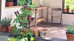 A covered patio area with a wooden table and chairs, surrounded by potted plants and a window with a view of greenery. - Typical cottage and outbuilding near Honfleur and Deauville  (QUETTEVILLE)