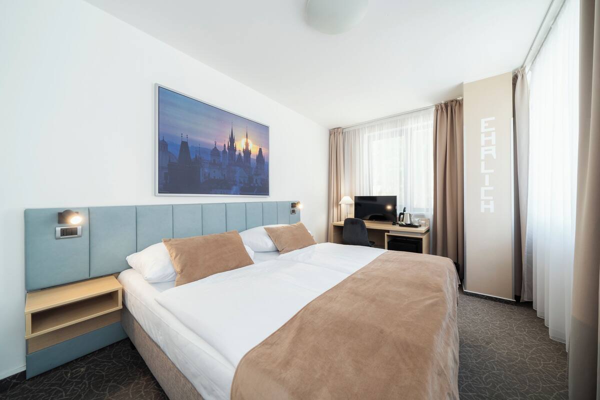 A hotel room with a large bed, a desk with a computer, a TV, and a cityscape painting on the wall.