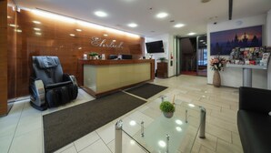 A reception area with a wooden reception desk, a massage chair, a glass table with a plant, and a wall-mounted TV. - Hotel Ehrlich Prague (Prague)