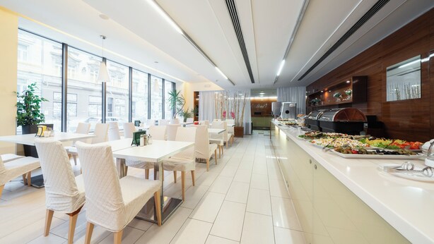 A modern dining area with white tables and chairs, a buffet counter with various food items, and large windows providing natural light. - Hotel Ehrlich Prague (Prague)