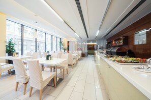A modern dining area with white tables and chairs, a buffet counter with various food items, and large windows providing natural light. - Hotel Ehrlich Prague (Prague)