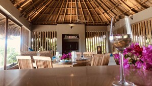 A dining area with a thatched roof, wooden furniture, and a glass of wine on the table. - TERRITORIO MAGICO (Dibulla)