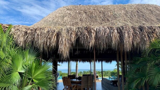 A covered outdoor area with a thatched roof, a dining table with chairs, and a view of the ocean. - TERRITORIO MAGICO (Dibulla)