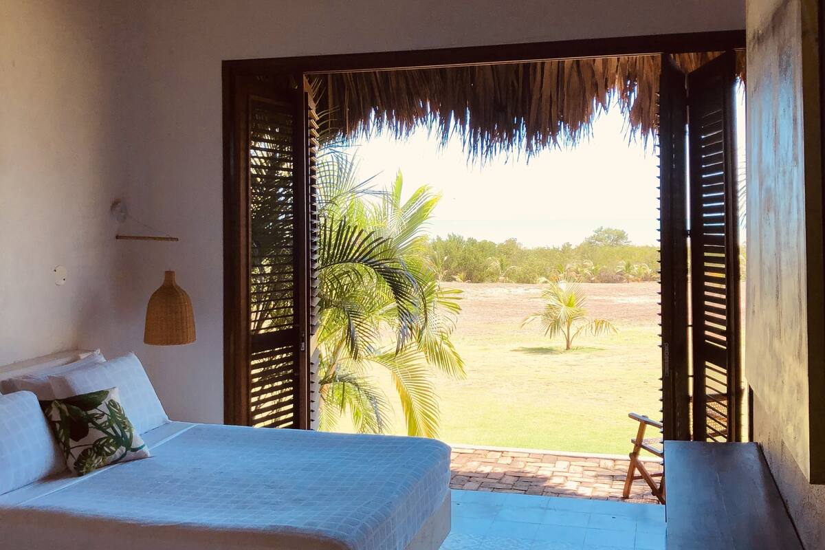A bedroom with a bed, a ceiling fan, a large window with a view of a grassy area and palm trees.