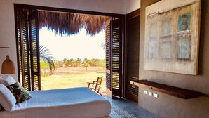 A bedroom with a bed, a chair, a painting, and a view of palm trees. - TERRITORIO MAGICO (Dibulla)