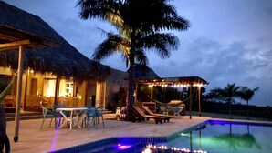 A pool area with lounge chairs and a palm tree, surrounded by a thatched-roof structure and a covered seating area with string lights. - TERRITORIO MAGICO (Dibulla)