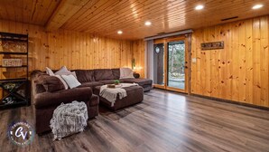 A wooden-paneled room with a brown sectional sofa, a small ottoman, and a glass door leading outside. - Family Cabin with hot tub on Cross lake (Crosslake)